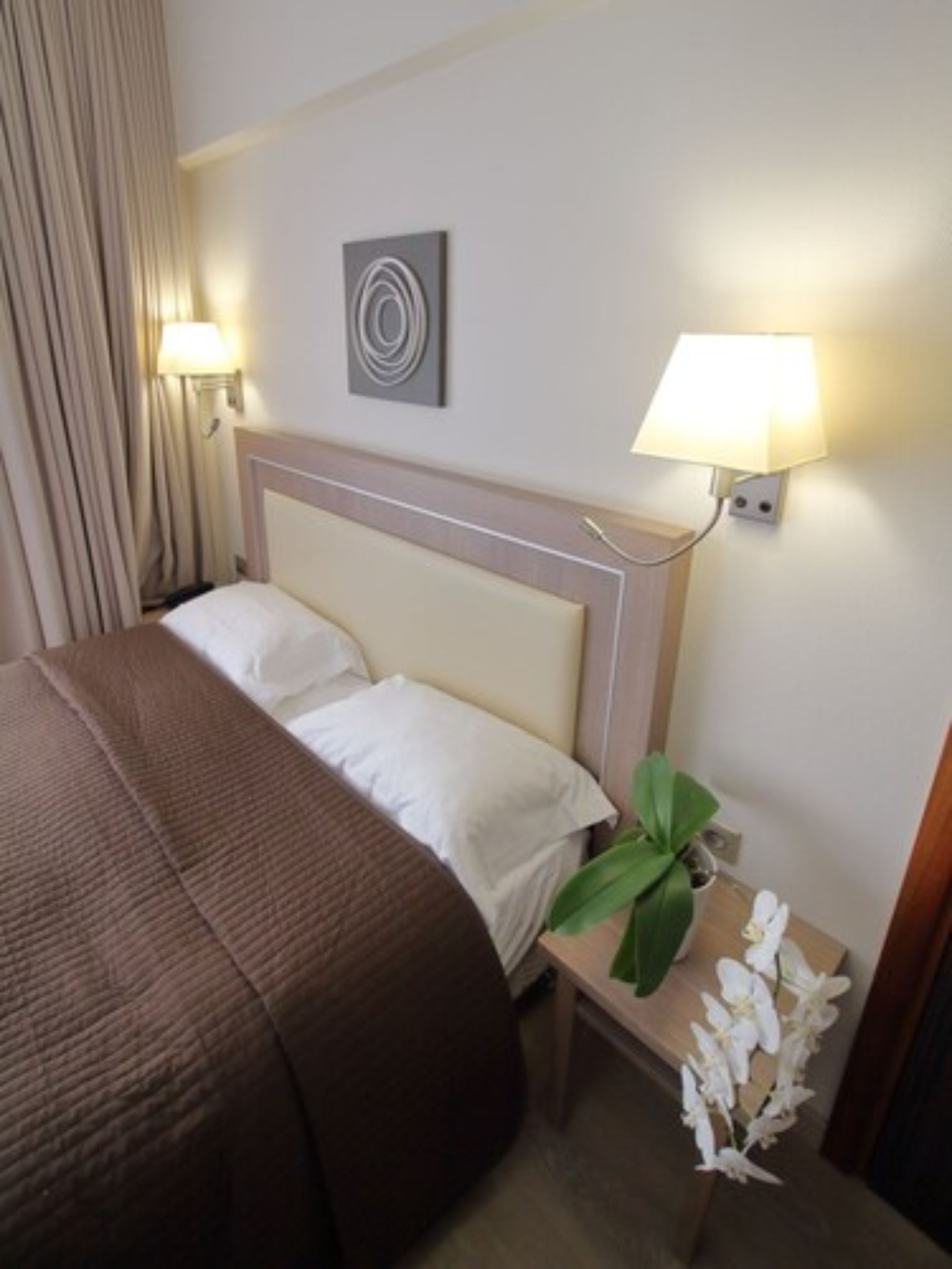 A hotel room with a bed, a nightstand with a lamp, a wall art piece, and a potted plant.