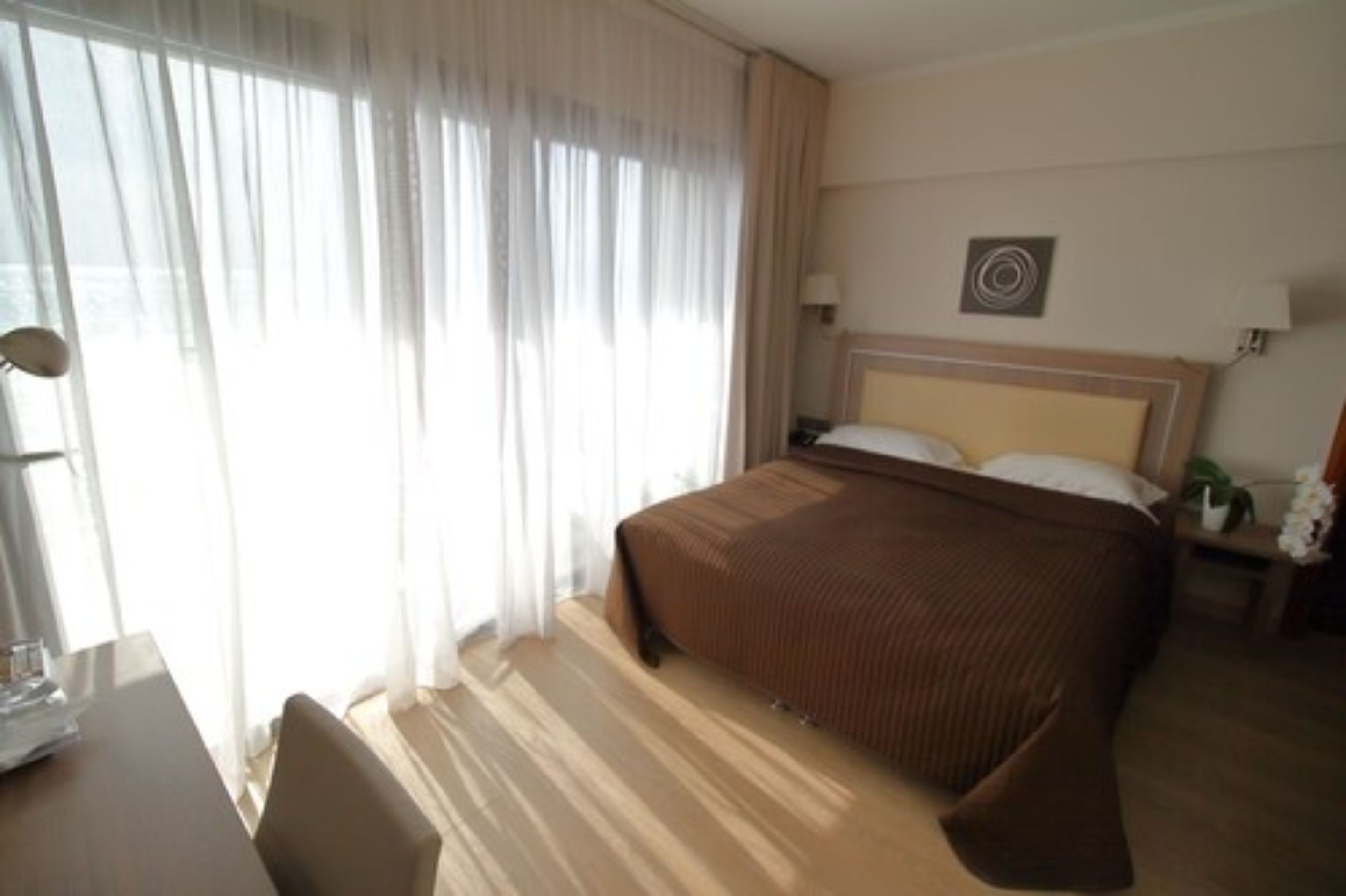 A hotel room with a large bed, a desk with a chair, and a window with sheer curtains.
