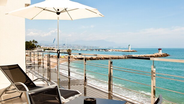 A balcony with a table and chairs, overlooking a coastal area with a pier and a lighthouse. - Hotel Villa Azur (Villeneuve-Loubet)