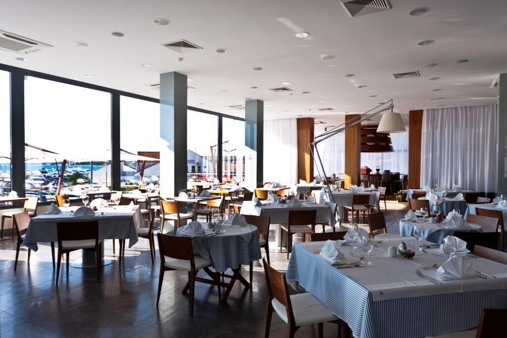 A spacious restaurant with high ceilings and large windows, offering views of the cityscape. Tables are neatly arranged with white tablecloths and set for dining.