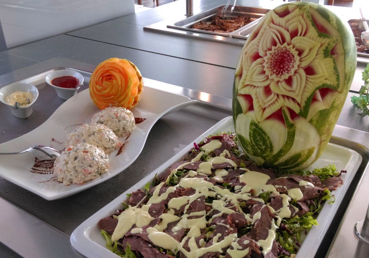 Een buffettafel met diverse gerechten, waaronder een creatief uitgesneden watermeloen, een salade met vlees en een bord met gehaktballetjes.