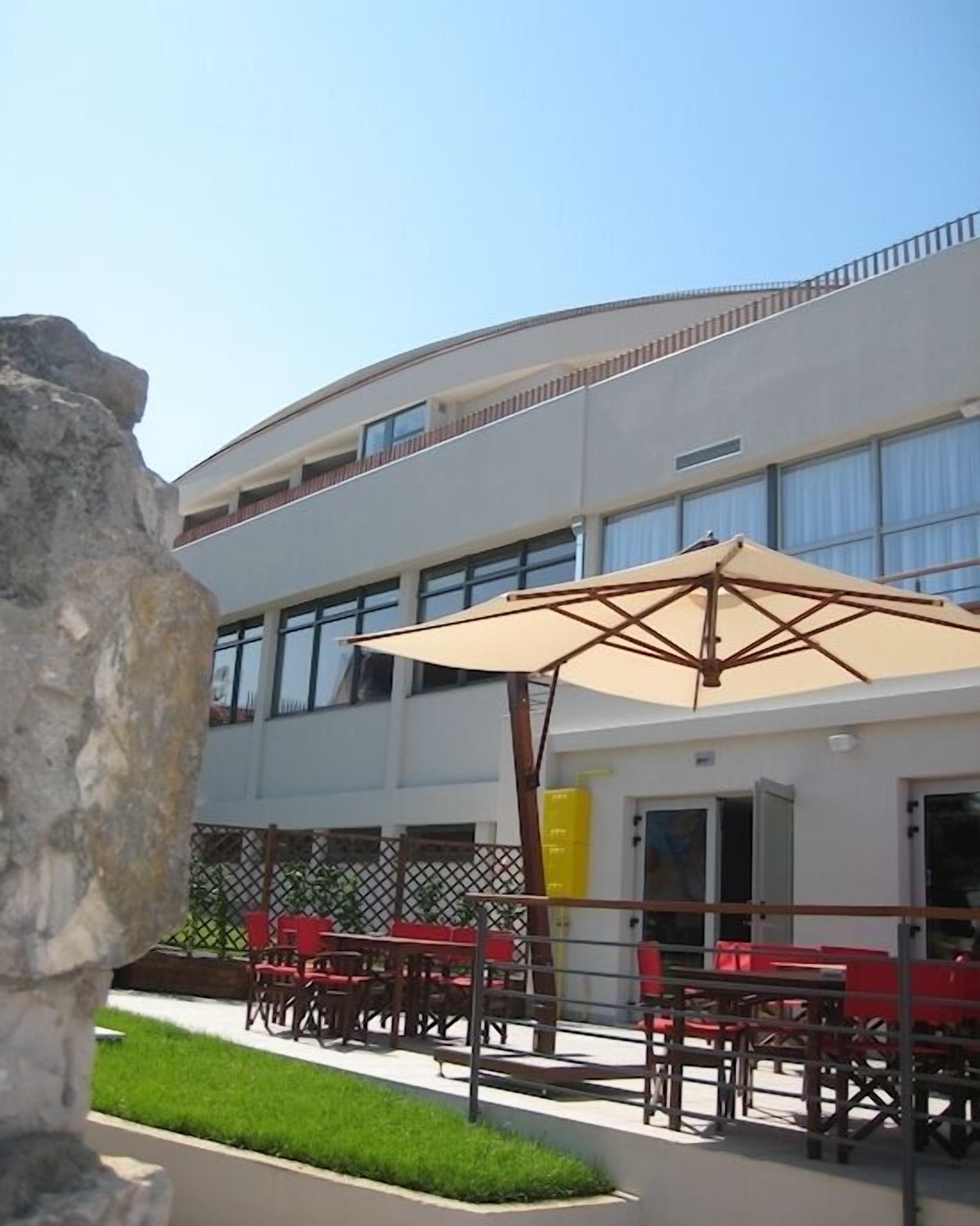 An outdoor seating area with tables and chairs, a large umbrella, and a building with multiple windows and balconies.