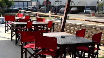 Mobiliario exterior con sillas y mesas rojas, un muro de piedra y un estacionamiento con coches y un edificio al fondo.
