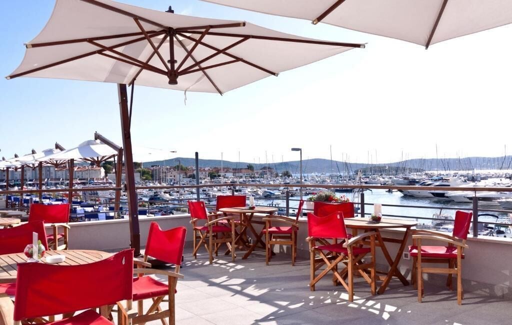 A terrace with red chairs and tables, white umbrellas, and a view of a marina with boats.