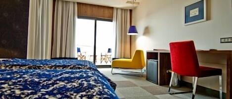 Ein Hotelzimmer mit einem Bett, einem Schreibtisch, einem roten Sessel, einer Lampe und Blick ins Freie.
