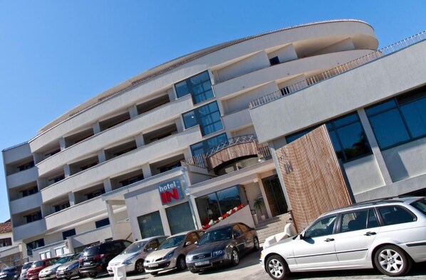 A multi-story hotel with a modern design, featuring balconies on the upper floors and a parking lot with several cars. - Hotel IN (Biograd na Moru)