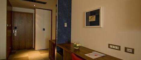 A hotel room with a wooden desk, a red chair, a small table with a plant, and a framed picture on the wall.