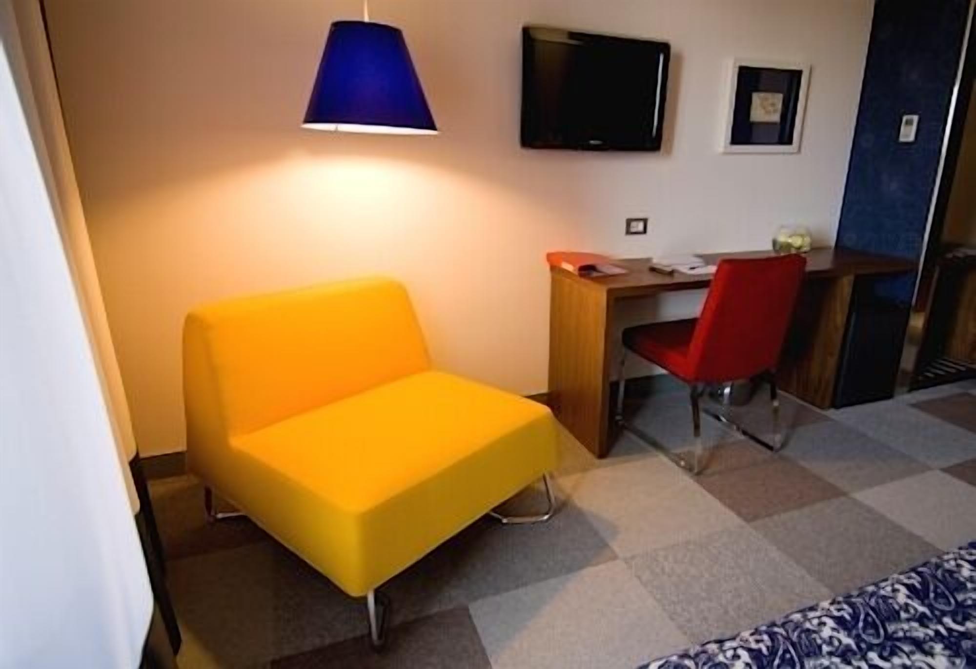 A room with a yellow chair, a desk with a red chair, a television, and a blue lamp.