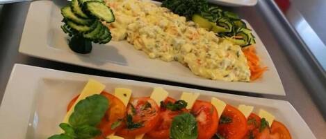 Un buffet proposant divers plats, notamment une salade composée de tomates et de fromage, un plat crémeux aux légumes, et une assiette de concombres et de carottes coupés en tranches.