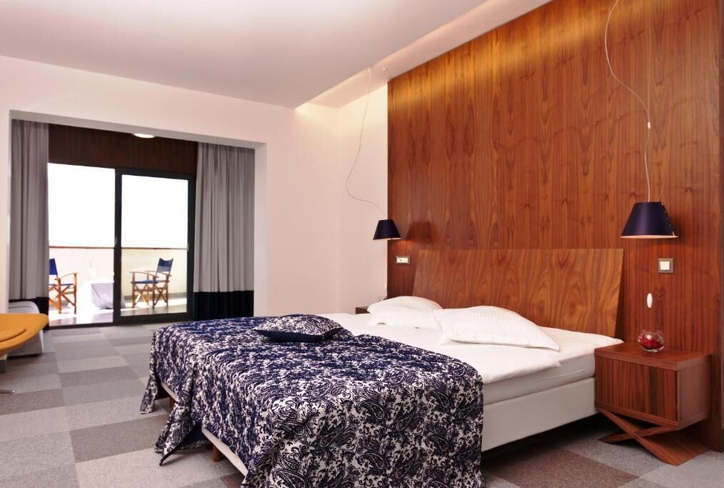 A modern hotel room with a large bed, a wooden accent wall, a small bedside table, and a view of the outdoor seating area.