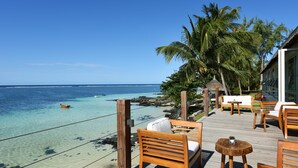 Une terrasse en bois, agréée de mobilier d’extérieur, qui offre une vue dégagée sur une mer d’un bleu profond, avec une structure à toit de chaume et des palmiers en arrière-plan.