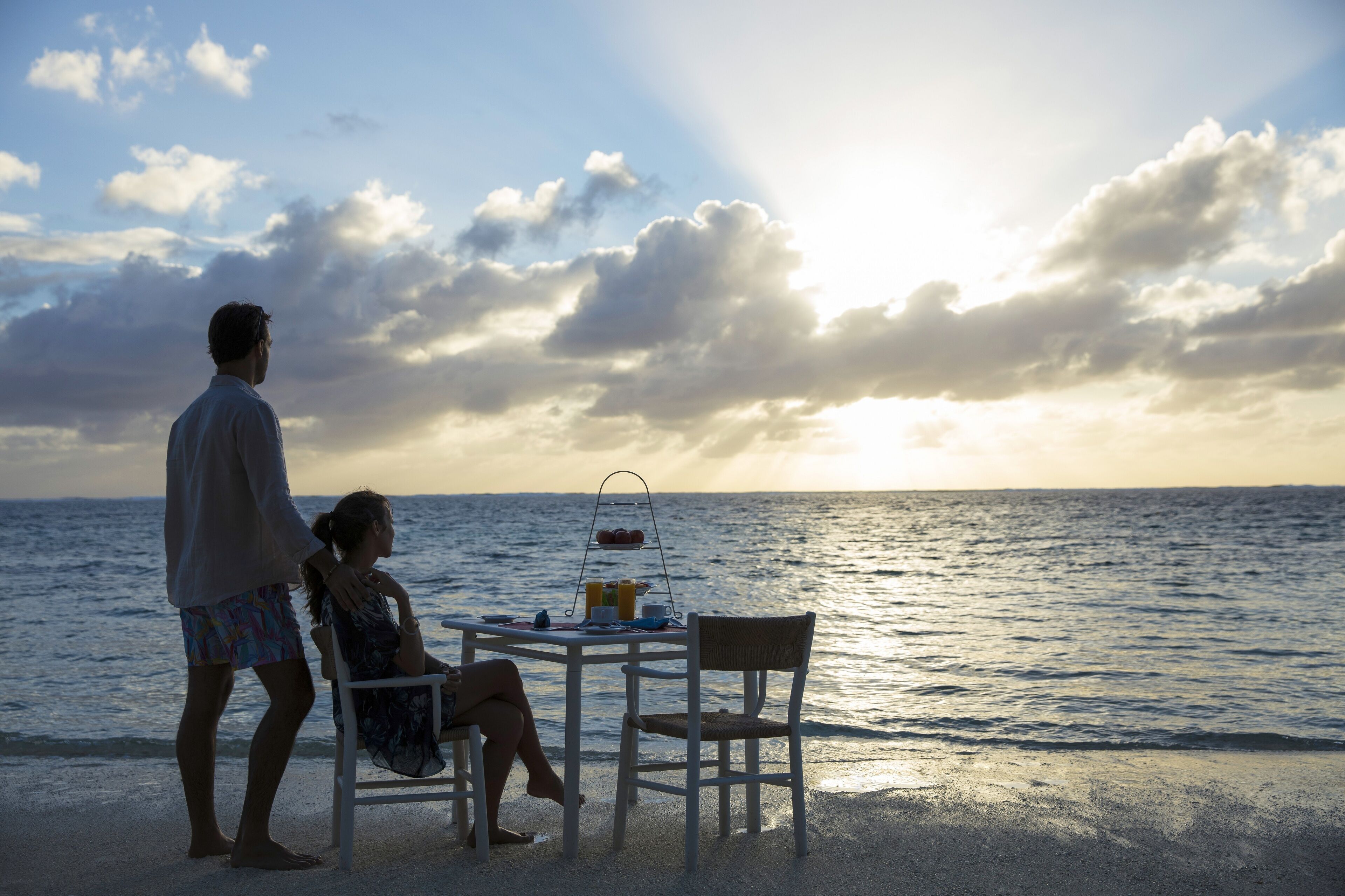 Escena de playa con una mesa y sillas, una persona de pie y un atardecer sobre el océano.