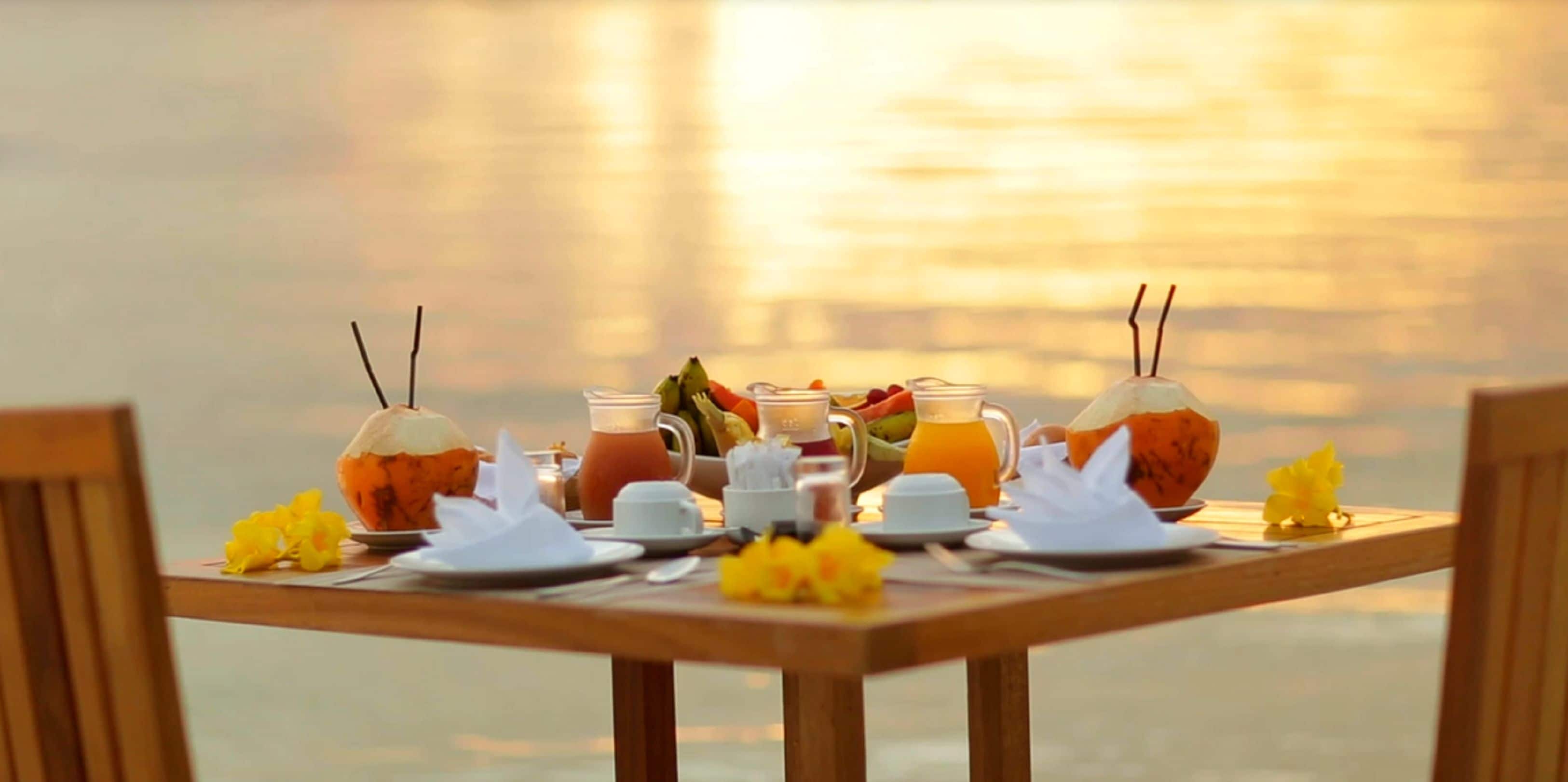 Una mesa de desayuno con diversas bebidas y frutas, con vista a un atardecer.