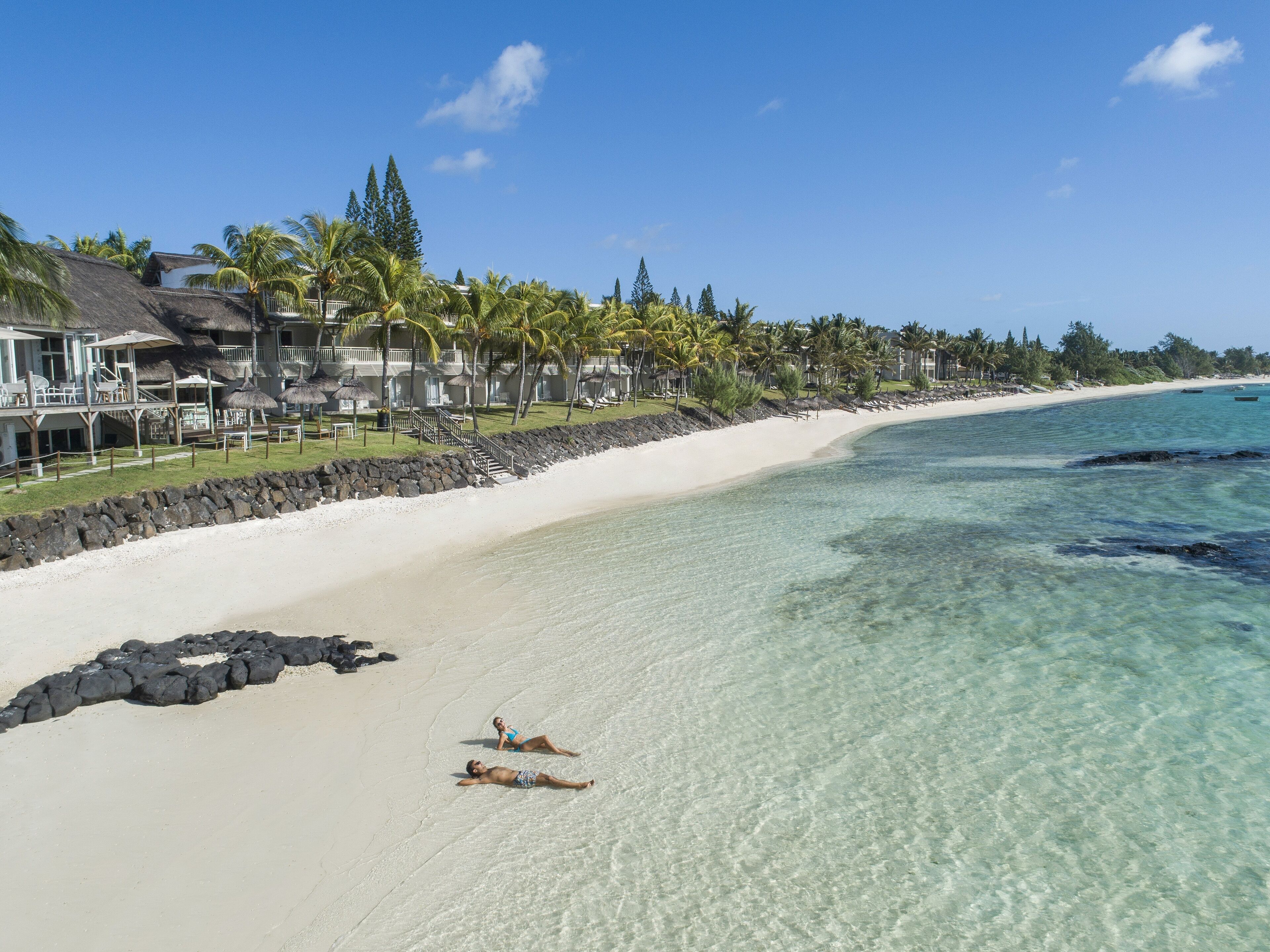 A beach with clear turquoise water, a white sandy shore, and a resort with thatched roofs and palm trees.