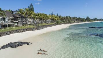 A beach with clear turquoise water, a white sandy shore, and a resort with thatched roofs and palm trees.