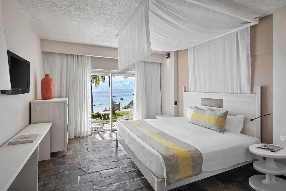 Habitación de hotel con cama, televisión, una mesita y vista al océano.