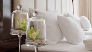 Un lit bien fait, avec du linge de lit blanc, une table de chevet en bois sur laquelle reposent deux vases à fleurs recouverts de verre, et une lampe fixée au mur.