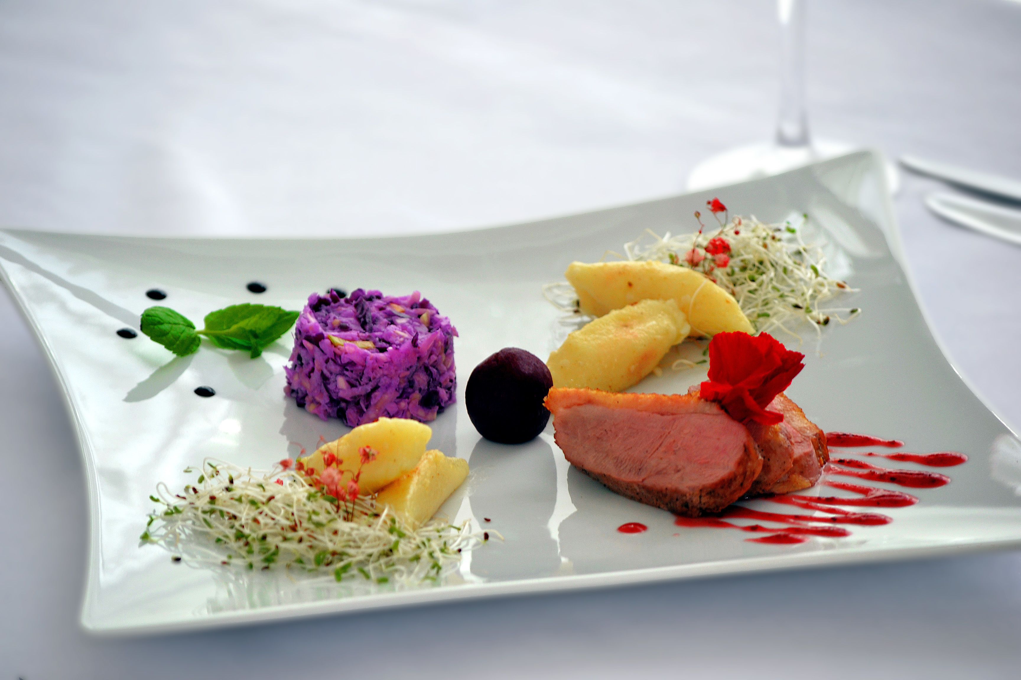 A square white plate with a variety of food items including purple-colored food, yellow food, a dark purple fruit, and a piece of meat with red sauce.