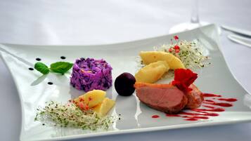 A square white plate with a variety of food items including purple-colored food, yellow food, a dark purple fruit, and a piece of meat with red sauce.
