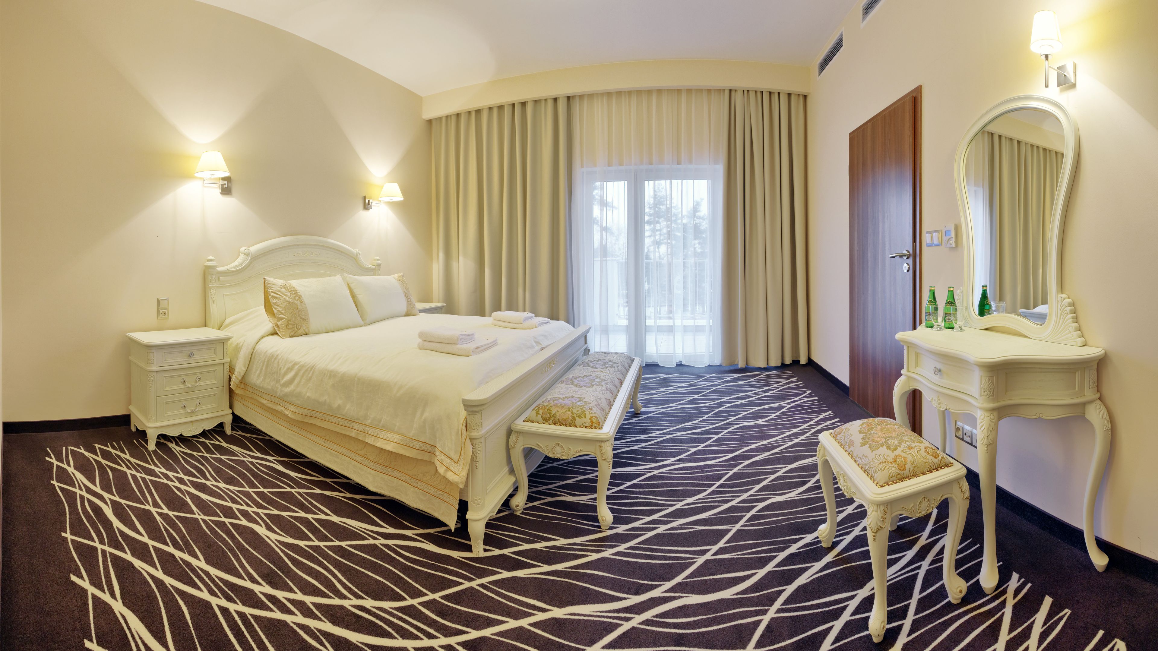 A hotel room with a large bed, a bedside table, a bench, a dressing table with a mirror, and a large window with curtains.