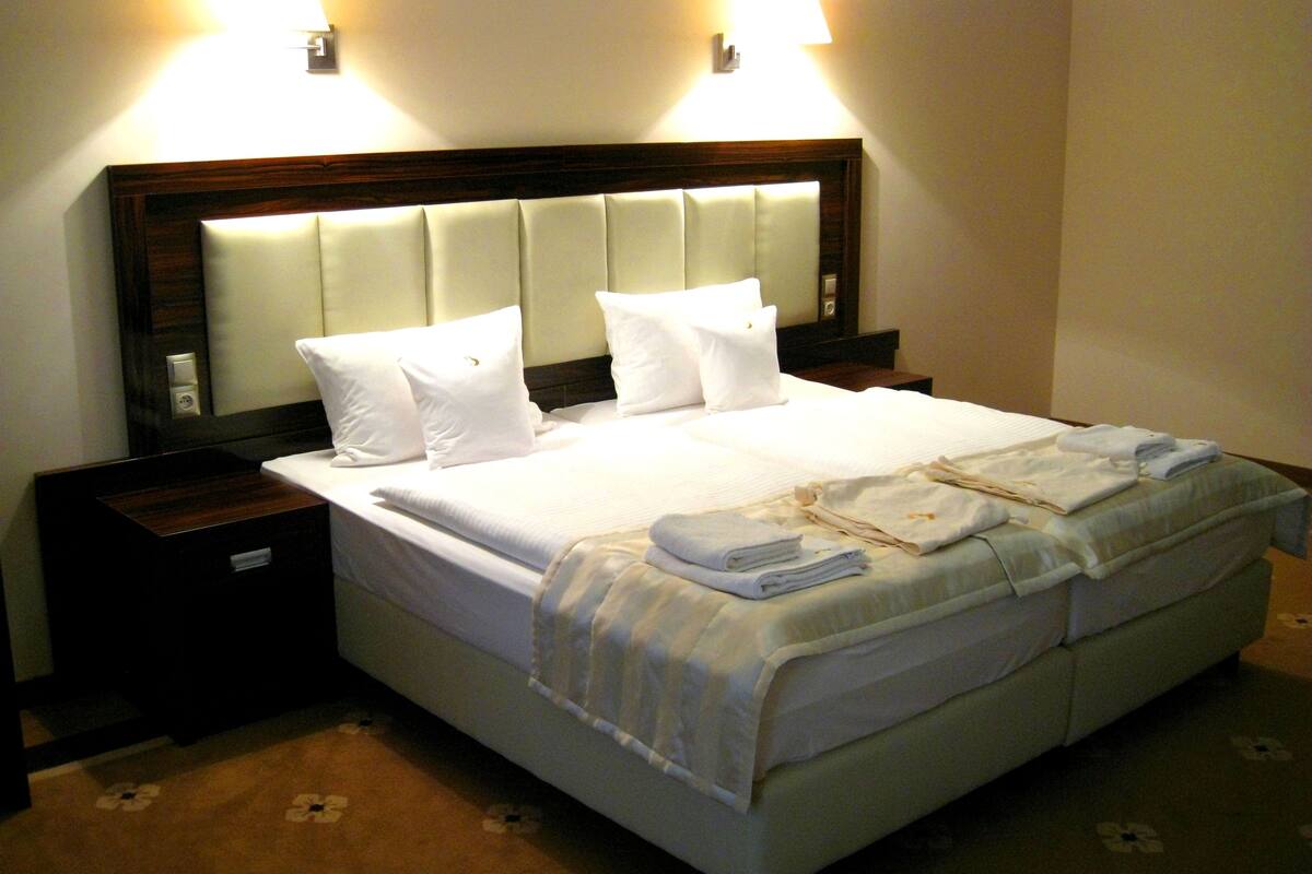 A neatly made bed with white linens and pillows, a bedside table with a lamp, and a floral carpet pattern.