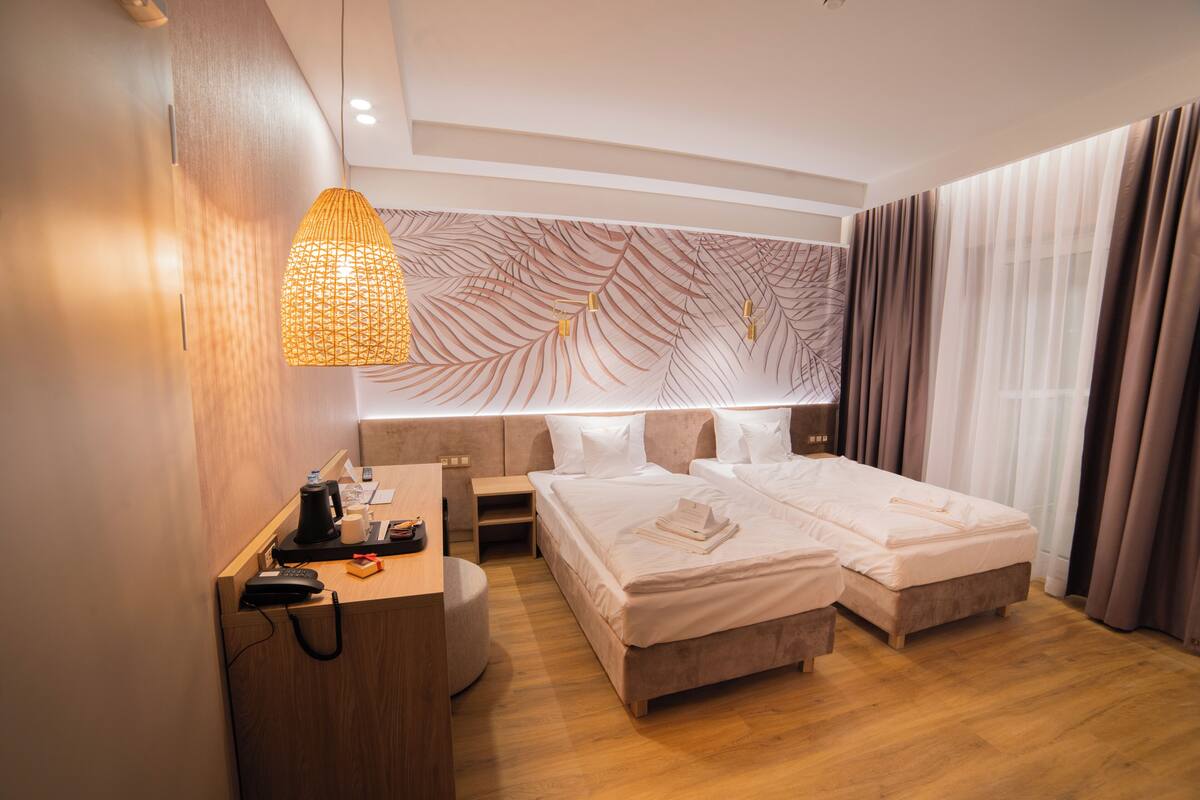 A hotel room with two beds, a desk with a telephone and a lamp, a wall with a leaf pattern, and a window with curtains.