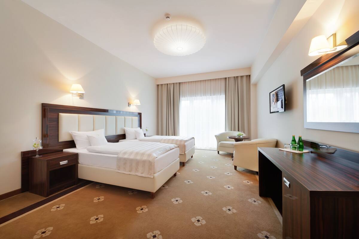 A hotel room with a bed, two bedside tables, a desk, a chair, and a large window with curtains.