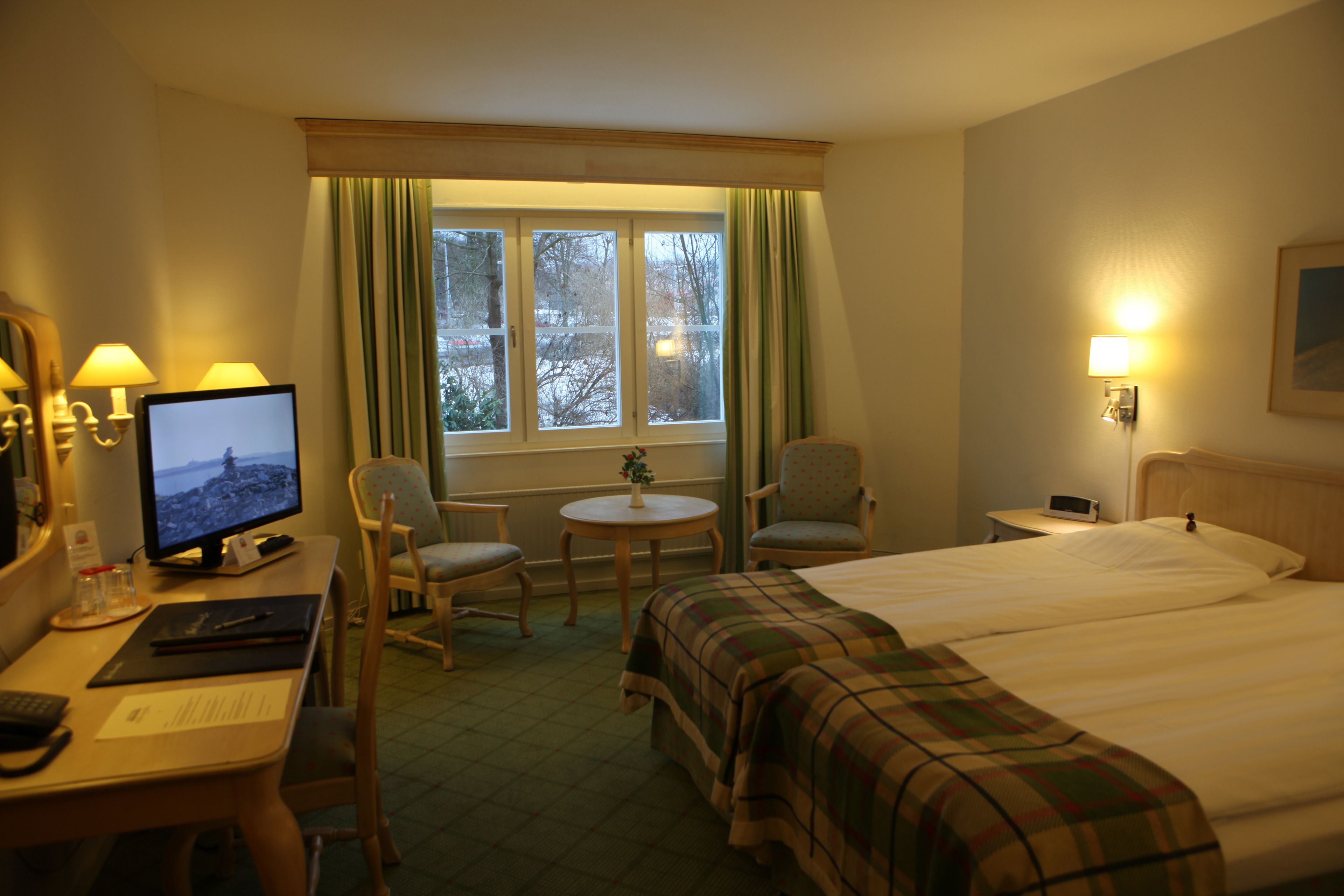 A hotel room with a bed, a desk with a computer, two chairs, a small table with a plant, and a window with curtains.