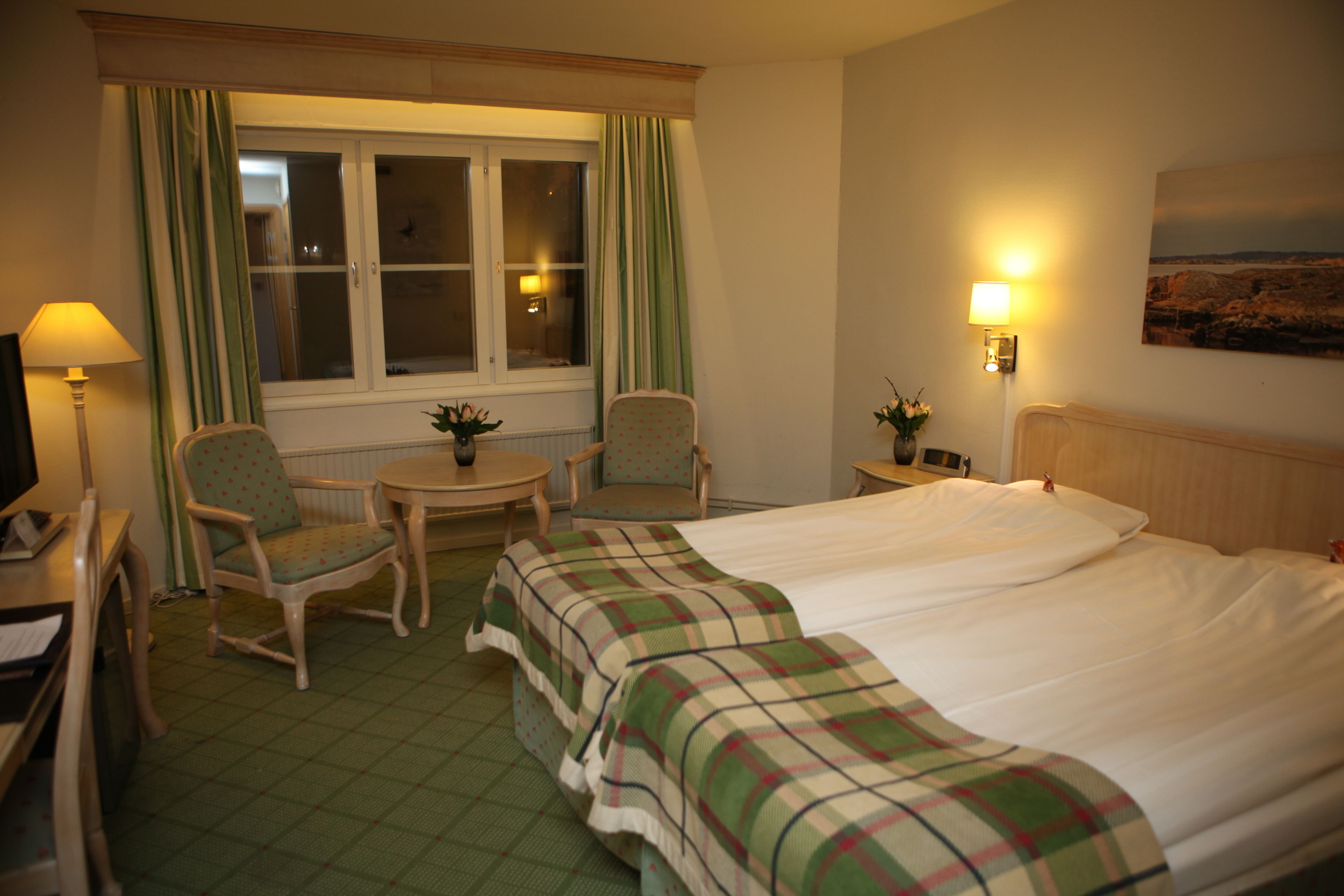 A hotel room with a bed, two chairs, a small table, a lamp, and a window with curtains.