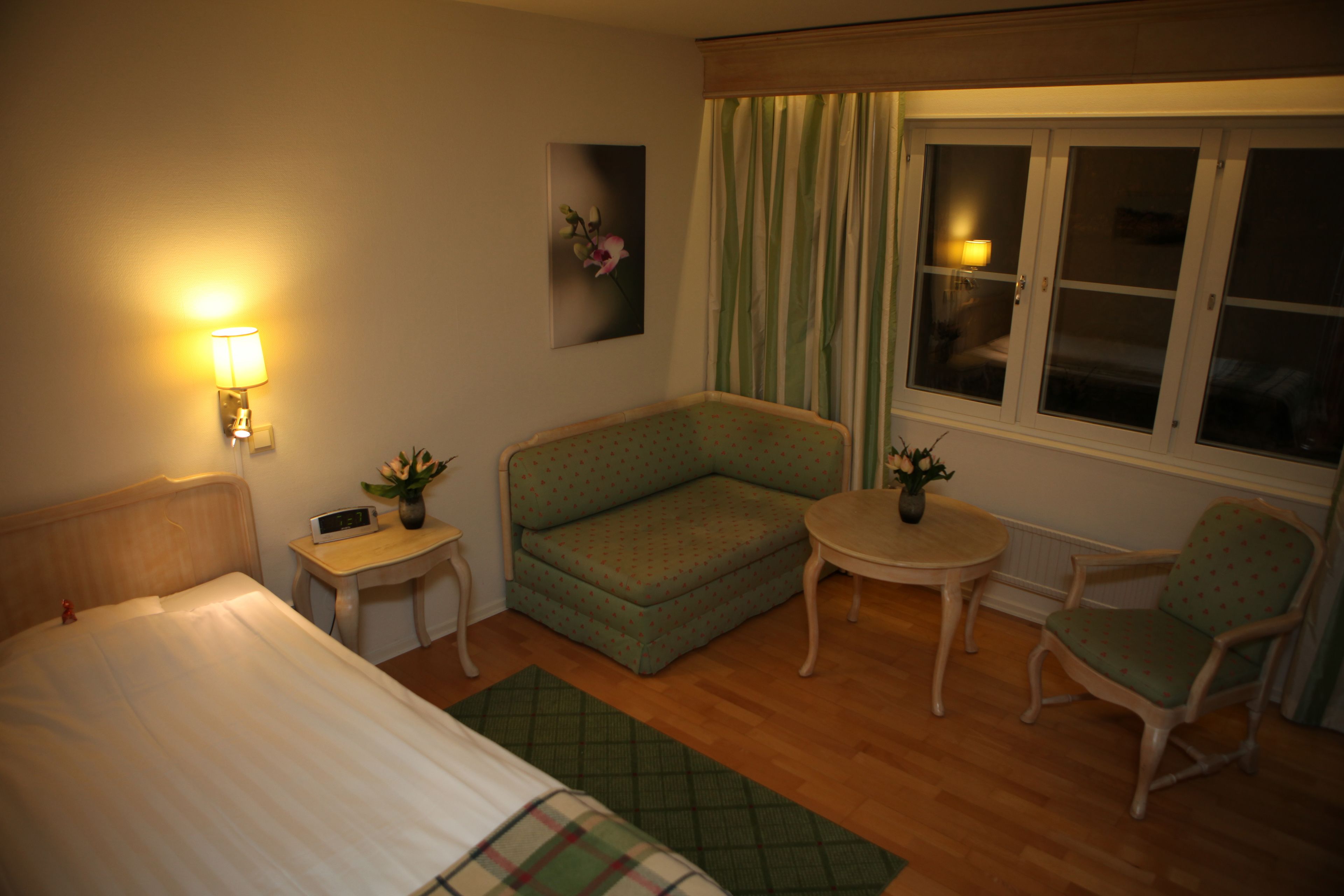 Ett hotellrum med en säng, en grön soffa, ett litet bord, en stol och ett fönster med gardiner.