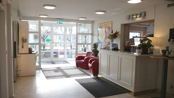 Un espace d’accueil doté d’une porte d’entrée en verre, d’un comptoir de réception, d’un coin salon avec des fauteuils rouges et d’une plante en pot.