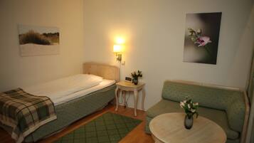 Una habitación pequeña y bien iluminada con una cama individual, un sofá verde, una mesita con un jarrón de flores y una lámpara de pared.