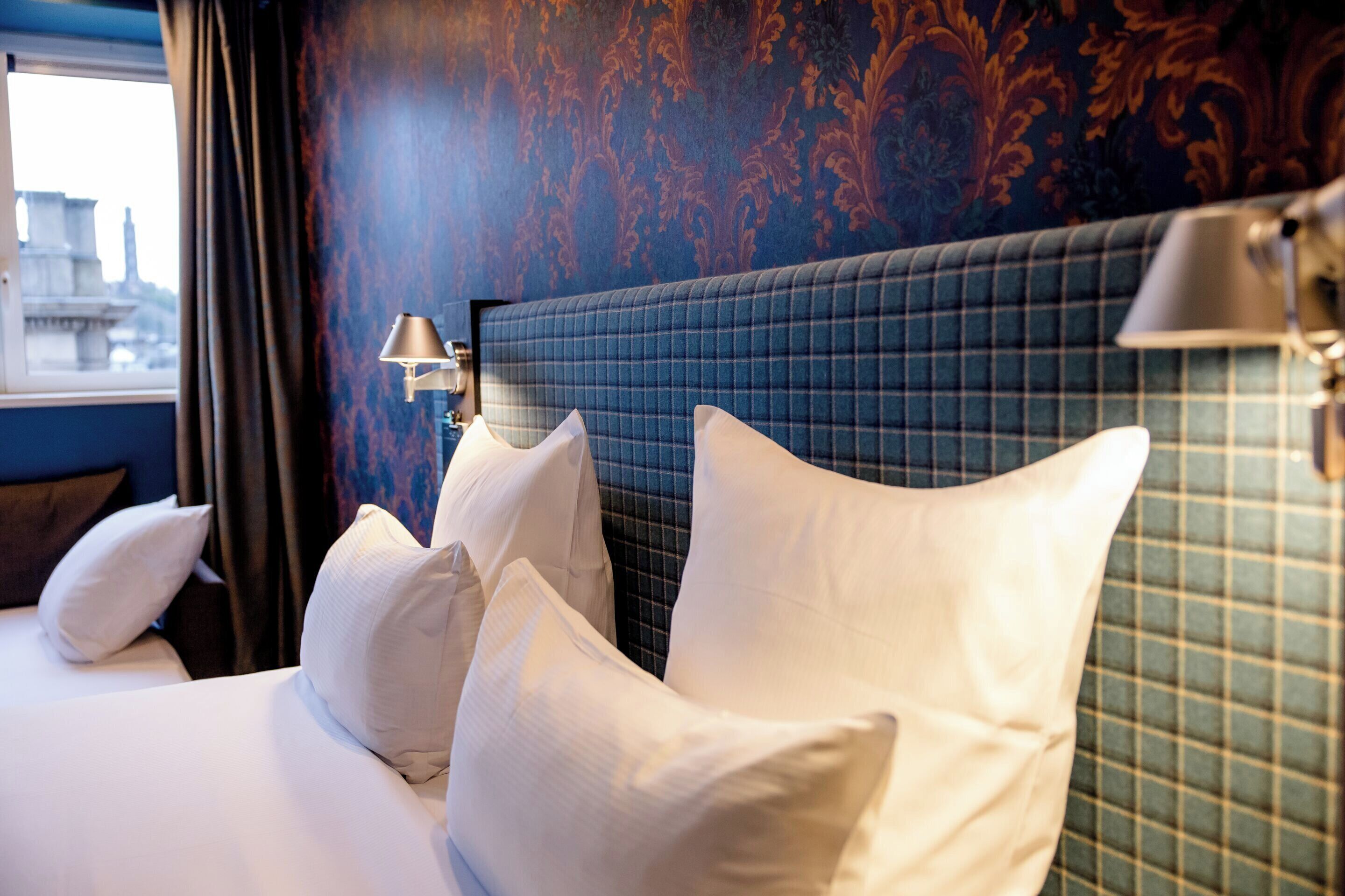 A hotel room with a large bed, multiple pillows, a headboard with a grid pattern, a wall lamp, and a window with curtains.
