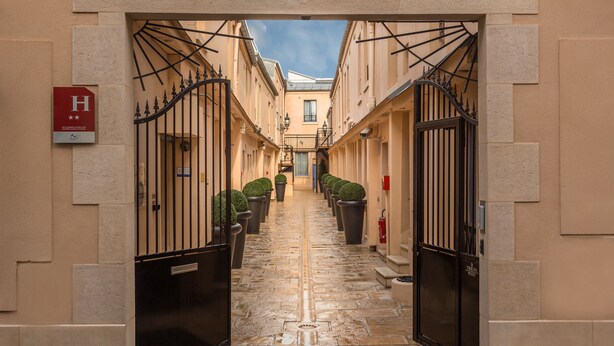 A narrow alleyway with a central pathway flanked by buildings, a double gate at the entrance, and a fire hydrant on the right. - Hôtel de L'Horloge (Paris)