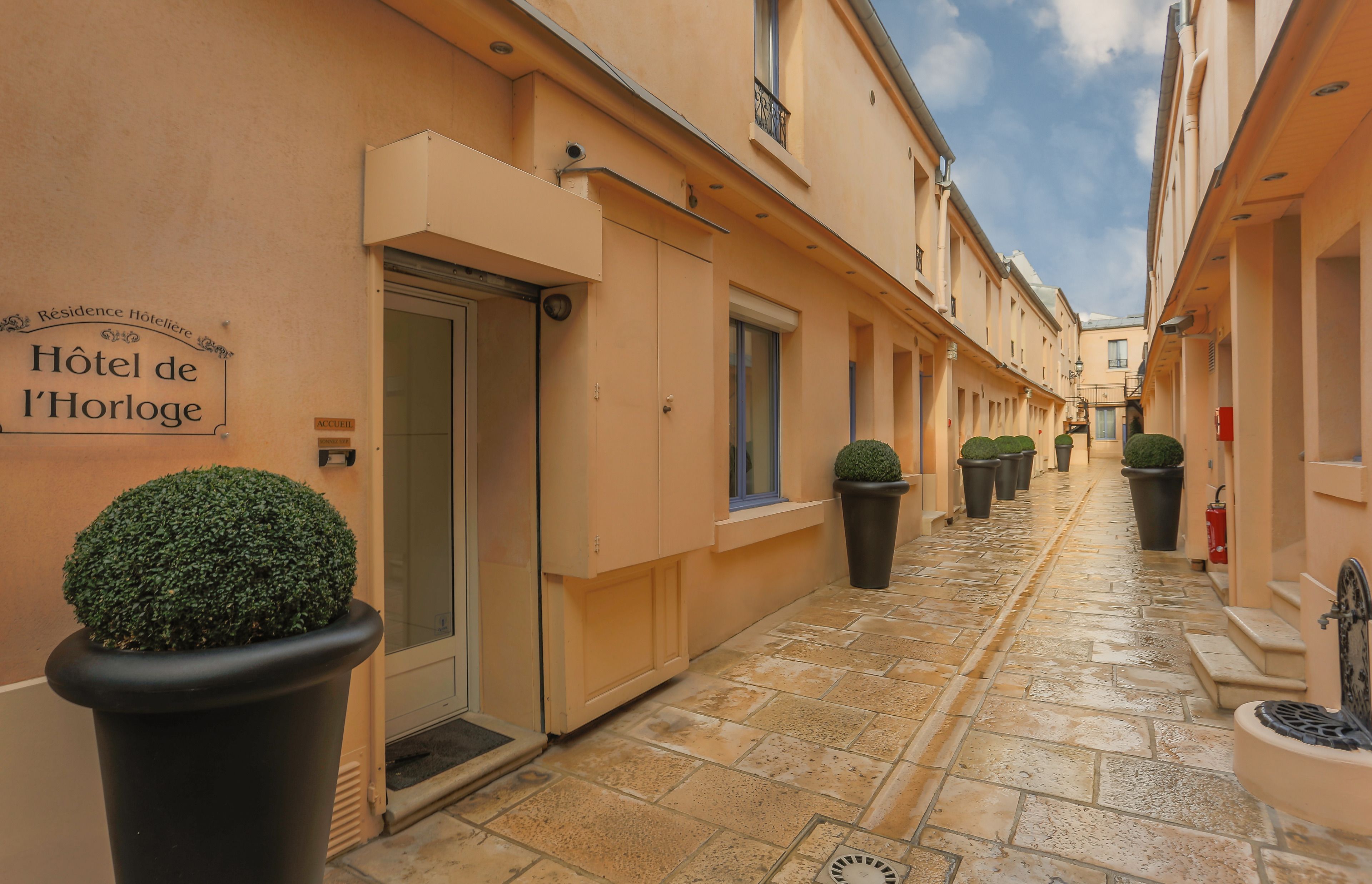 A narrow, paved alleyway with a row of buildings on either side, each with a door and windows. There are potted plants along the walkway.