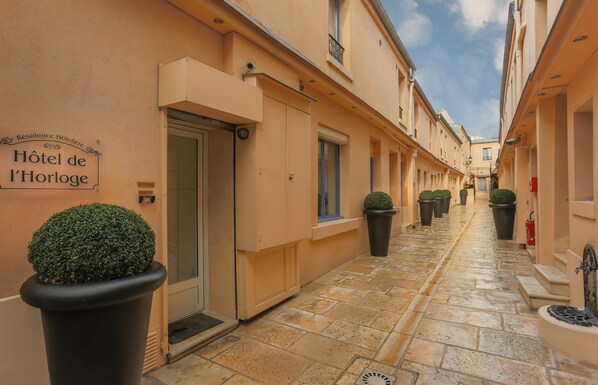 A narrow, paved alleyway with a row of buildings on either side, each with a door and windows. There are potted plants along the walkway.