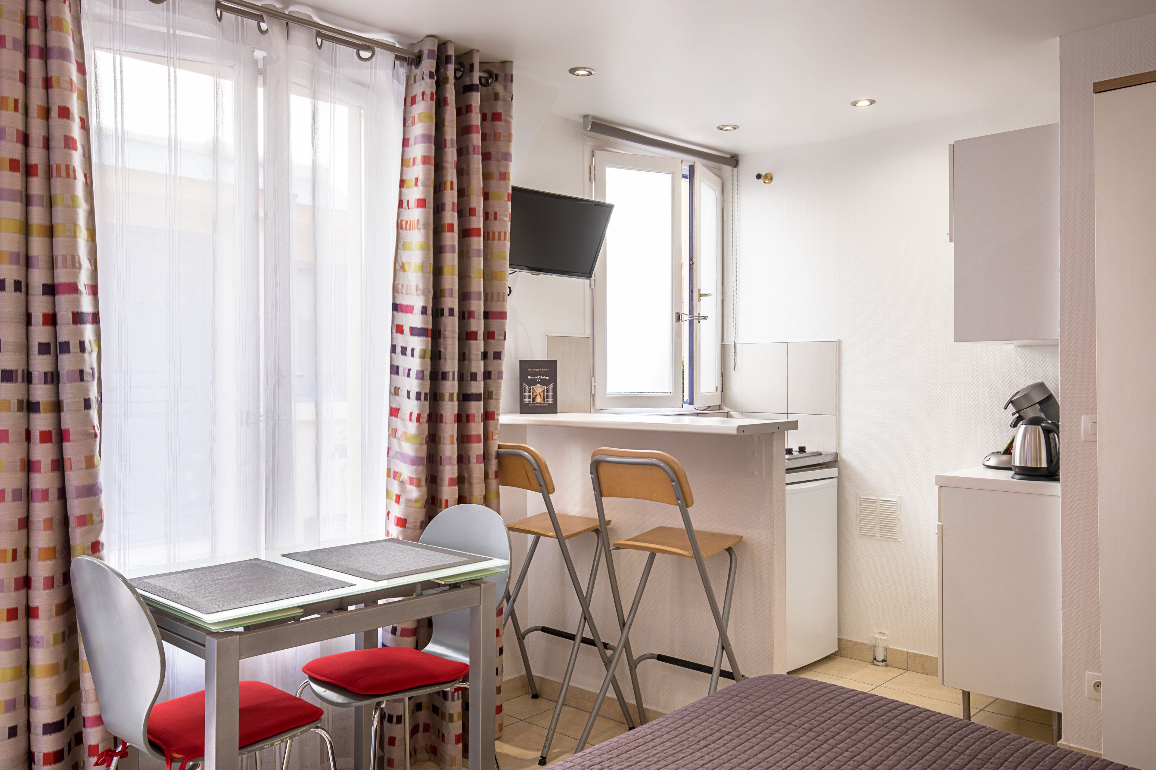 Una camera d'albergo compatta con un tavolo da pranzo, due sedie, un letto, angolo cottura e TV a parete.