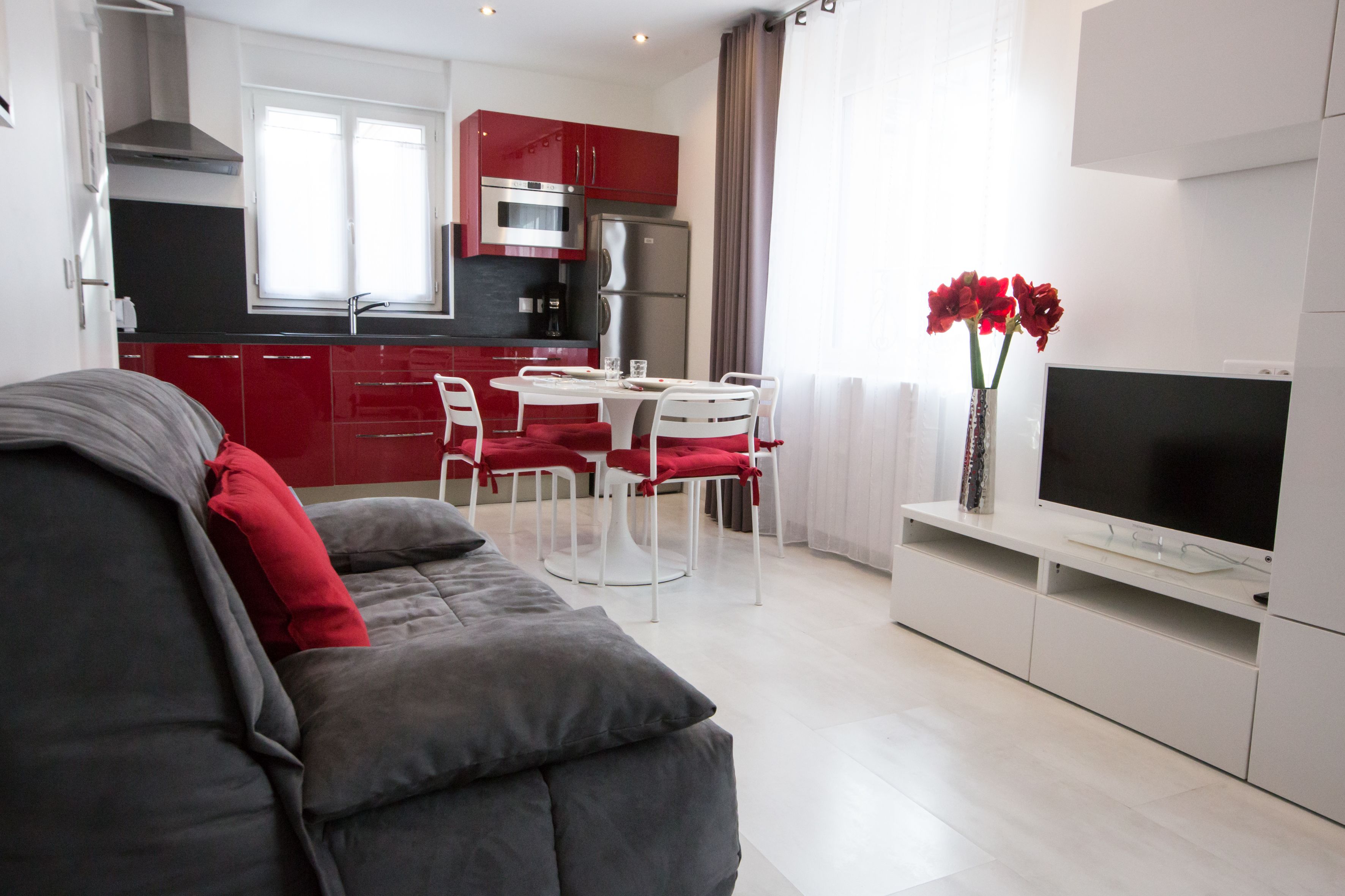 A modern living room with a grey sofa, red cushions, a dining area with white chairs, a TV on a white stand, and a kitchen with red cabinets and stainless steel appliances.