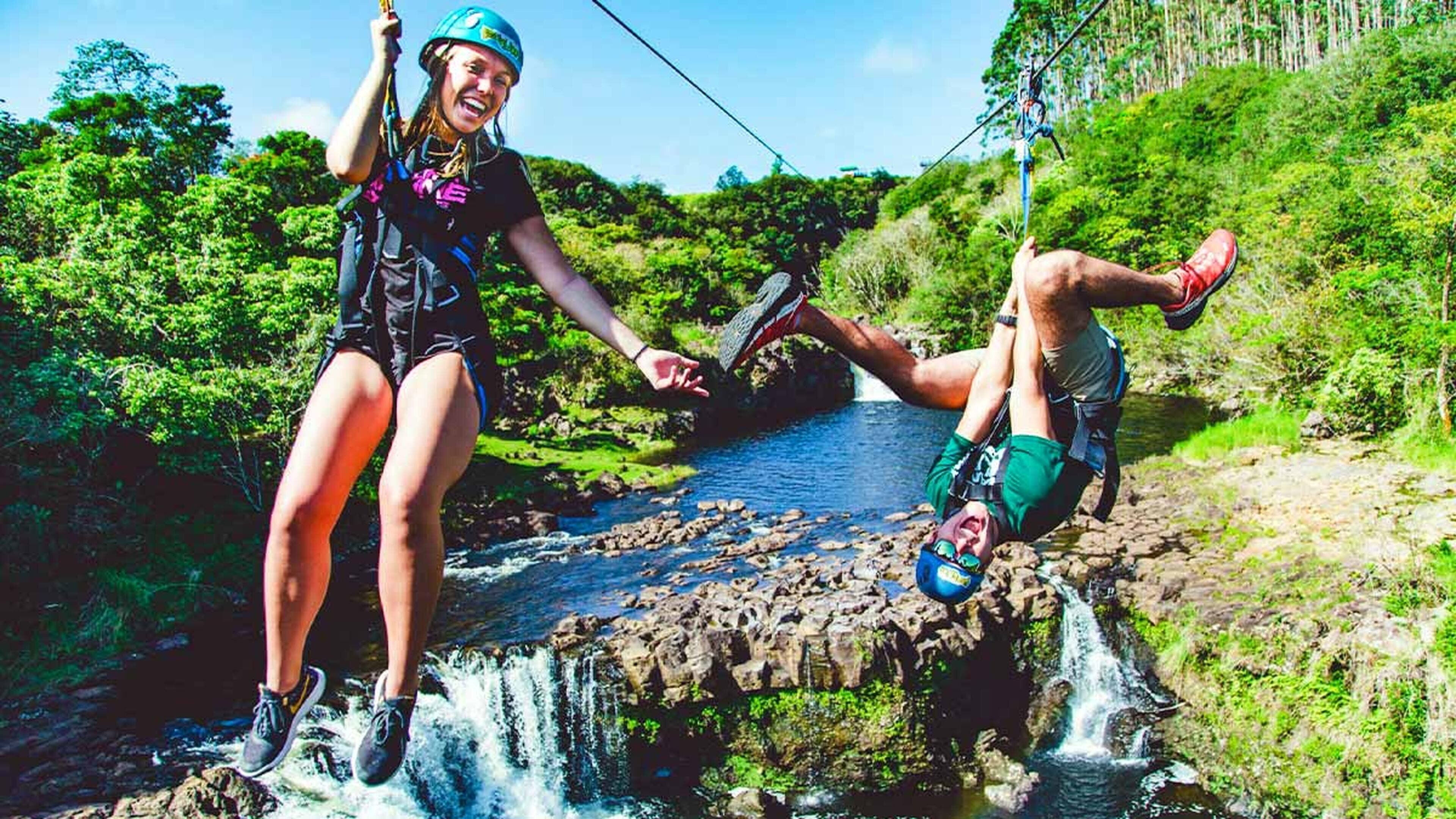 Dos personas realizan una actividad al aire libre cerca de un río con una cascada, usando cascos y arneses.