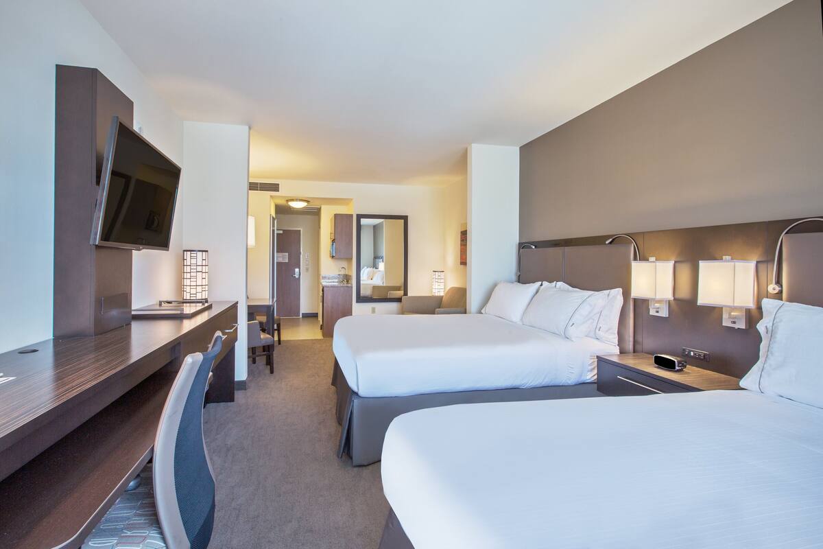Habitación de hotel con dos camas, un escritorio, un televisor y un baño con espejo e iluminación.