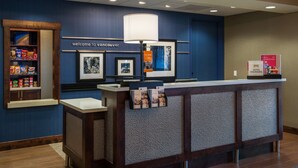 A reception desk with a 'Welcome to Vancouver' sign, a lamp, framed pictures, and a snack shelf. - Hampton Inn & Suites Portland/Vancouver (Vancouver)
