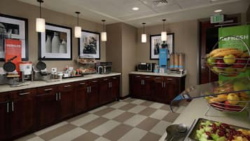 Una cocina de hotel con piso a cuadros, armarios de madera, una estación de café y una exhibición de frutas.