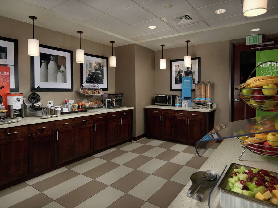 Una cocina de hotel con piso a cuadros, armarios de madera, una estación de café y una exhibición de frutas.