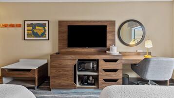Habitación de hotel con una mesita de madera para televisor, un televisor de pantalla plana, un espejo, un escritorio con lámpara y una cama.