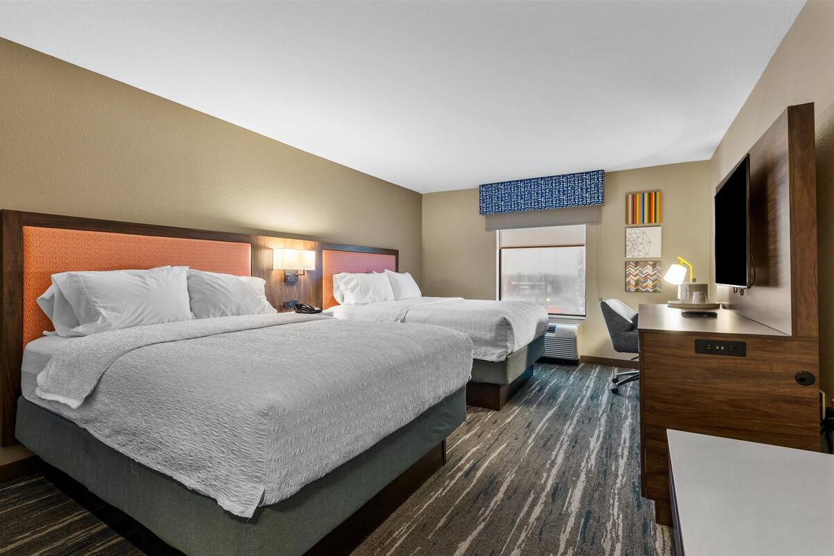 Quarto de hotel com duas camas, uma mesa, uma televisão e uma janela.