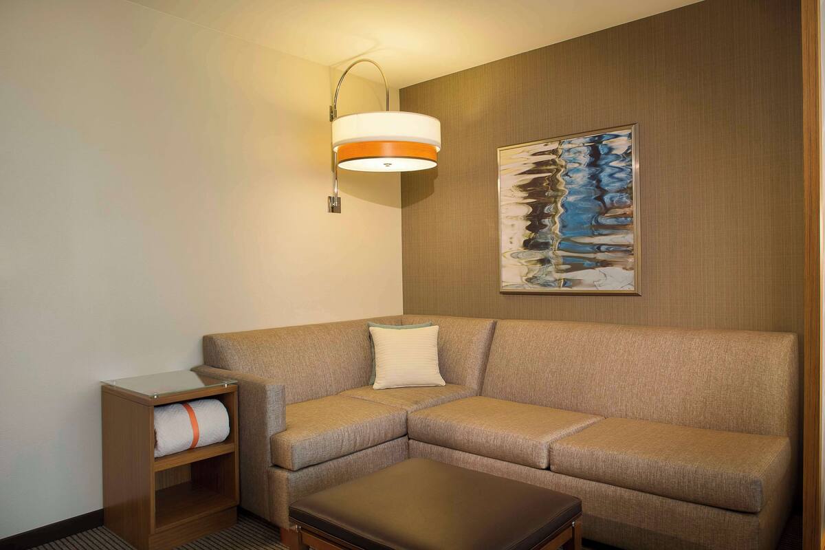A modern hotel room with a sectional sofa, a small wooden side table, a wall-mounted lamp, and a framed abstract painting.