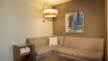 Quarto de hotel moderno com sofá de canto, uma pequena mesa lateral de madeira, luminária de parede e uma pintura abstrata emoldurada.