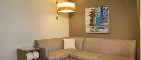 A modern hotel room with a sectional sofa, a small wooden side table, a wall-mounted lamp, and a framed abstract painting.
