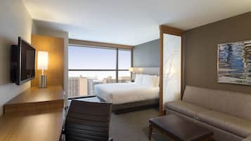 Quarto de hotel com cama, sofá, escrivaninha, cadeira, TV e uma grande janela com vista para a cidade.