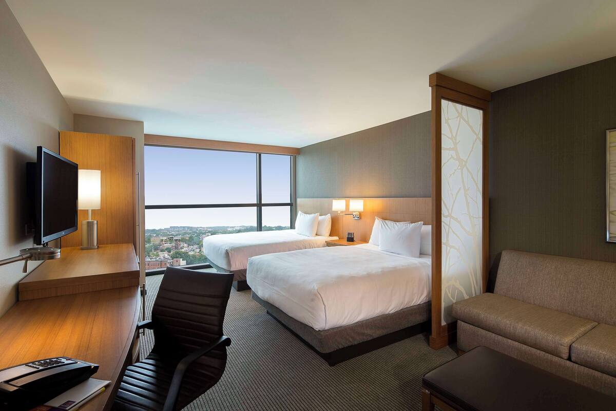 A hotel room with two beds, a desk with a TV, a sofa, and a large window offering a city view.